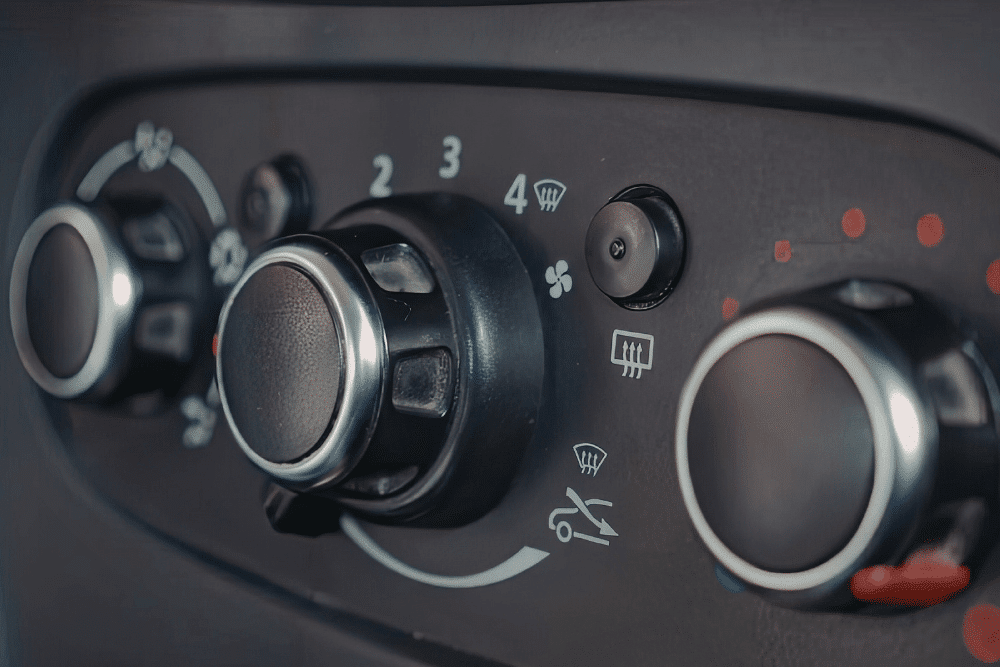 Close-up of a cars climate control panel, showing knobs for temperature, fan speed, and airflow direction, with icons for air conditioning, defrost, and air recirculation.