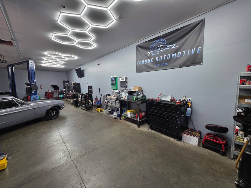 Auto repair in Madison, WI at Torque Automotive. Automotive workshop with tools, equipment, and storage shelves along the wall. A gray car is parked on the left. A large Torque Automotive banner hangs on the wall. The ceiling has hexagonal light fixtures.