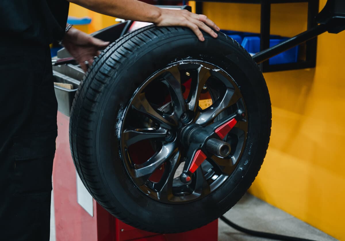A person holds a car tire and wheel mounted on a balancing machine in a workshop with yellow walls and brake repair equipment in the background.