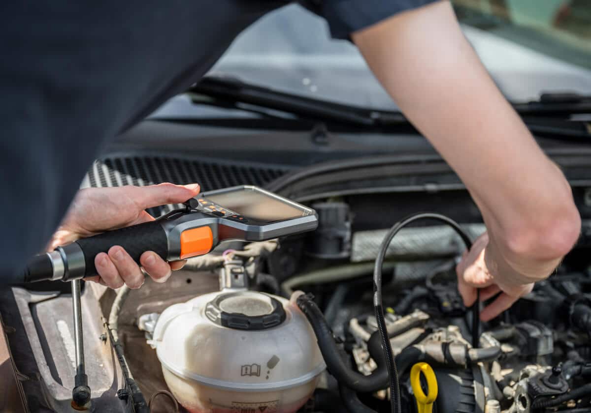A mechanic uses a diagnostic tool to inspect a car engine, holding the device in one hand while adjusting engine components with the other.