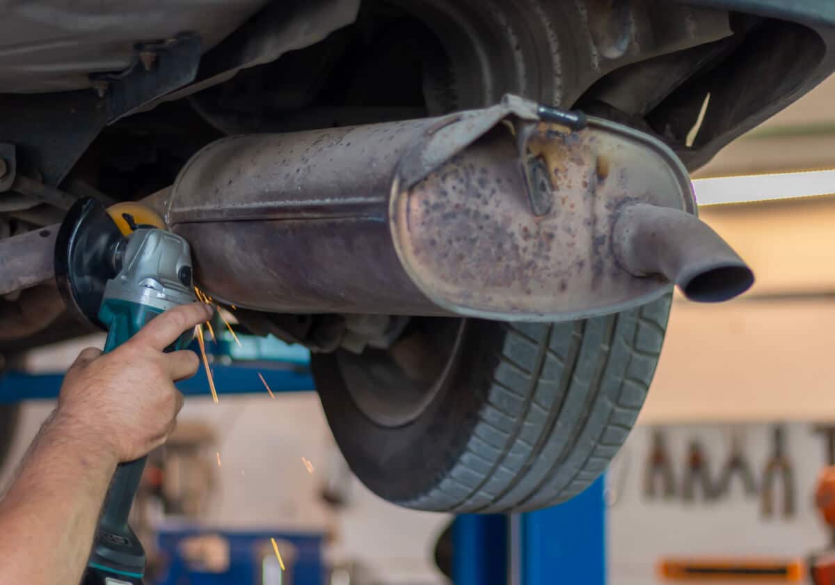 A person uses an angle grinder to cut a cars exhaust pipe near the muffler, with sparks flying, under a vehicle on a lift in a garage.