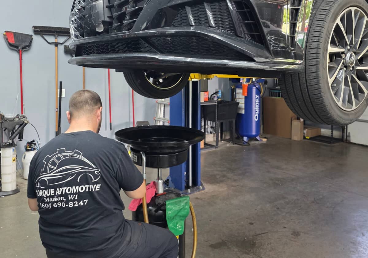 A mechanic performs an oil change under a lifted car in a garage. The mechanic wears a black Torque Automotive shirt, and tools are visible on the wall behind him.