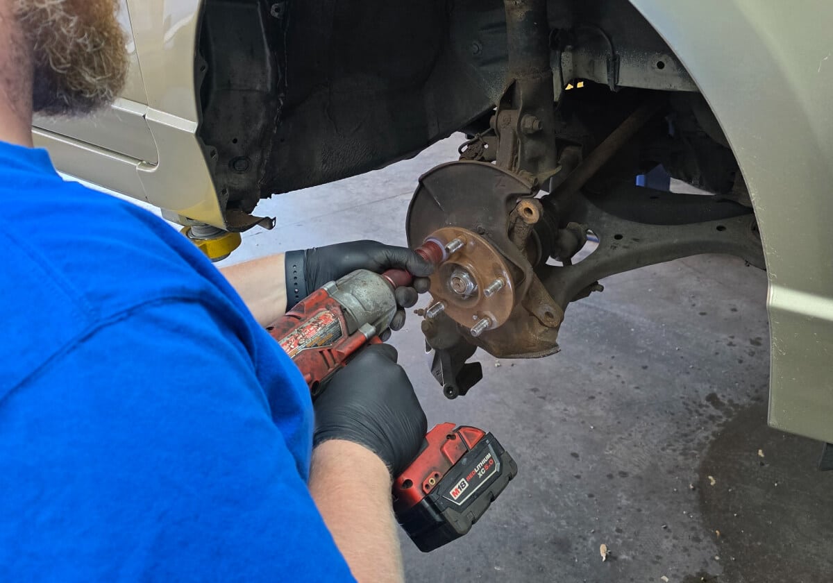 A person in black gloves uses a red power tool to work on the exposed brake assembly of a vehicle with the wheel removed.
