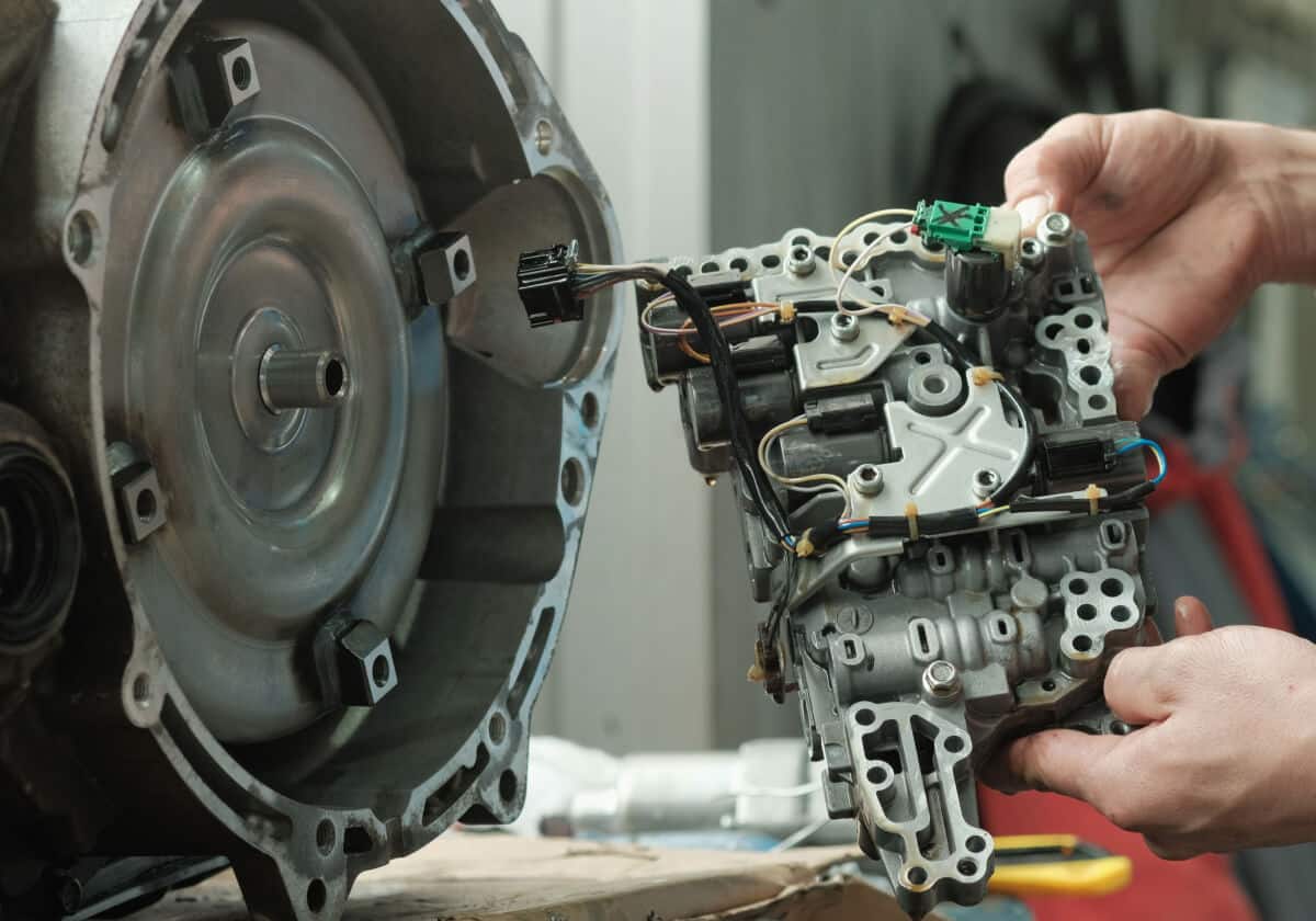 A person holds a disassembled automatic transmission valve body next to a transmission housing on a workbench.